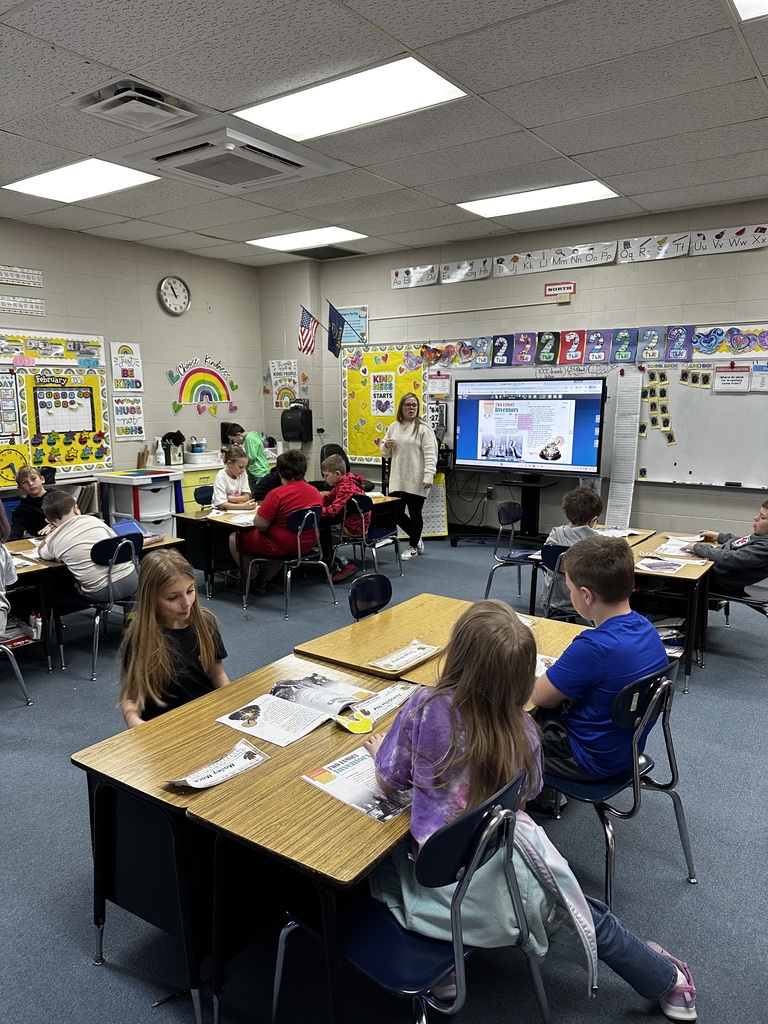 A classroom with students seated at desks. A teacher stands near a screen. Papers and books are on the desks.
