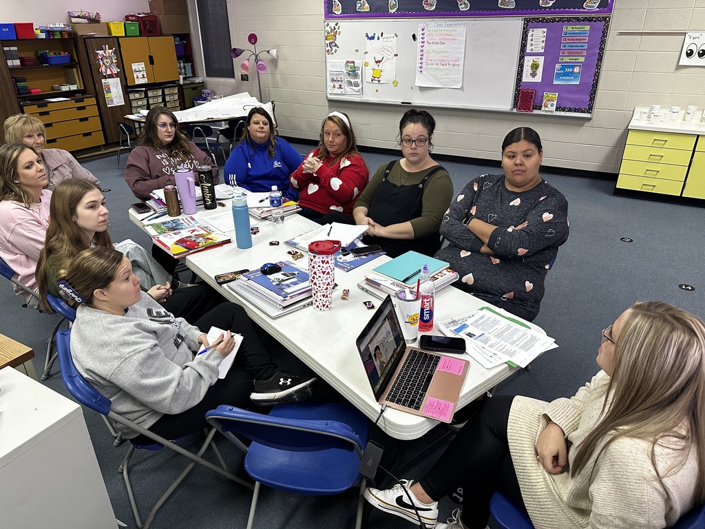 A group of people seated around a table in a classroom, some with laptops, books, and papers.