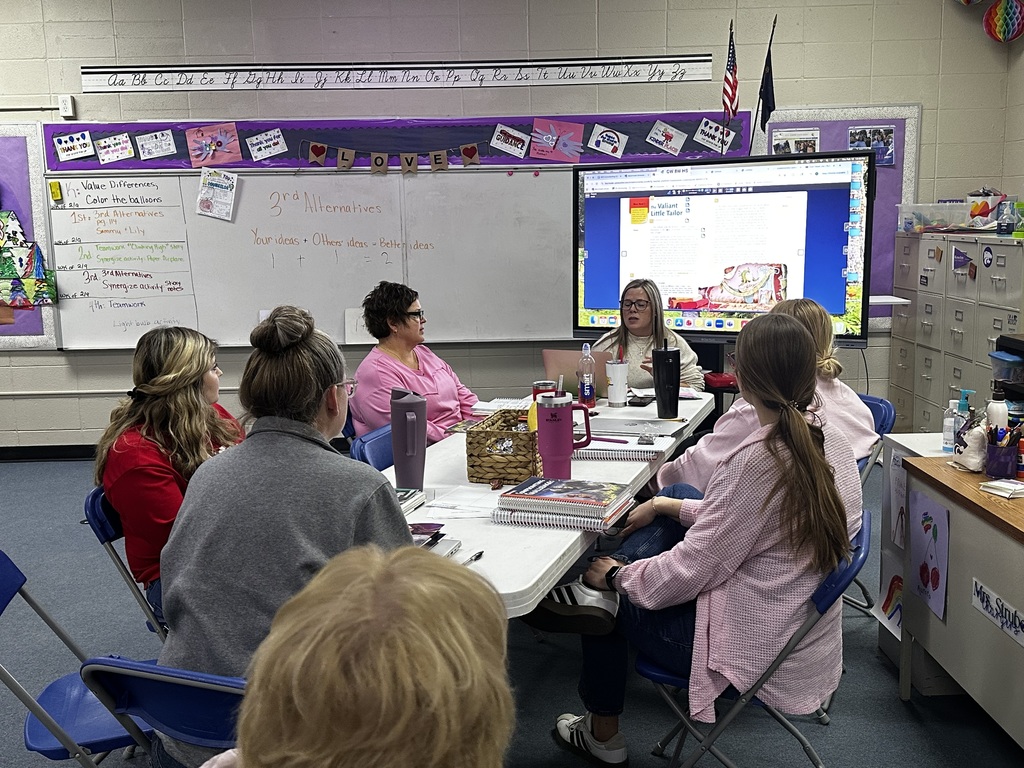 A group of people in a classroom, seated around a table, with a whiteboard and projector screen in the background.