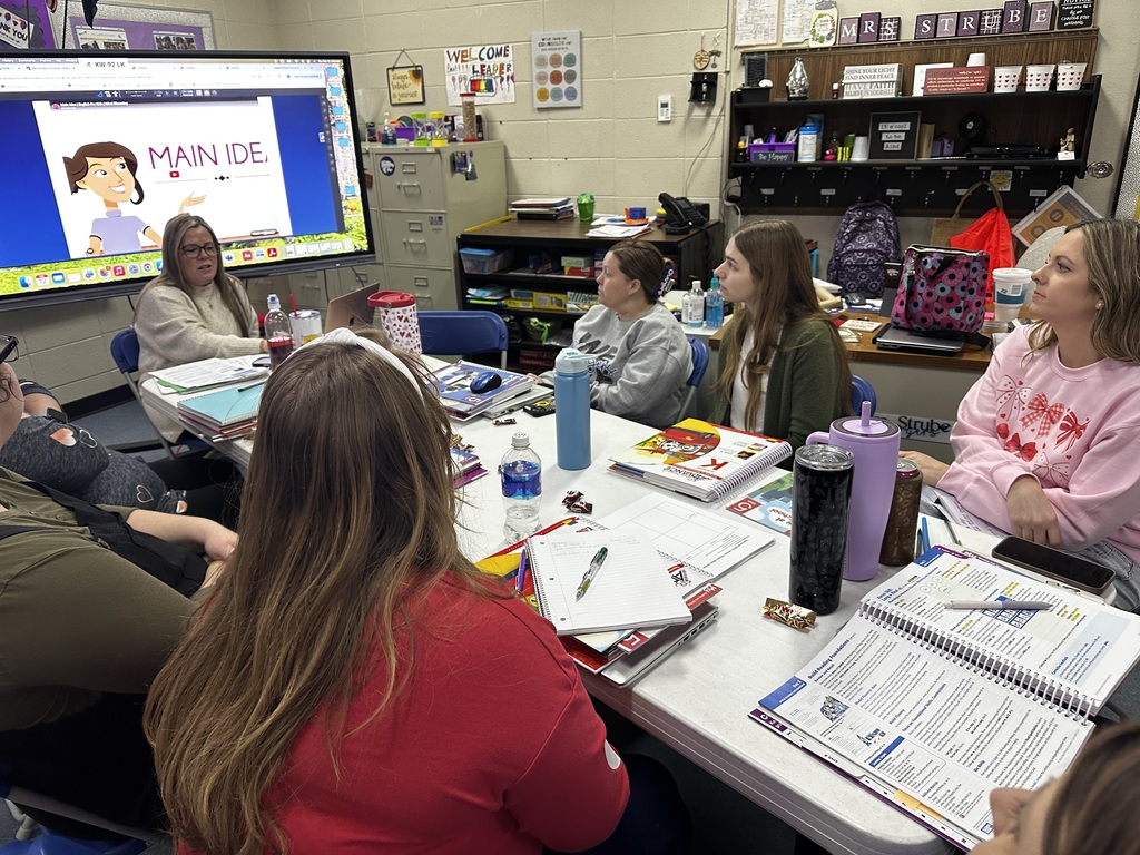 A group of people sit around a table with notebooks and pens, facing a classroom screen.