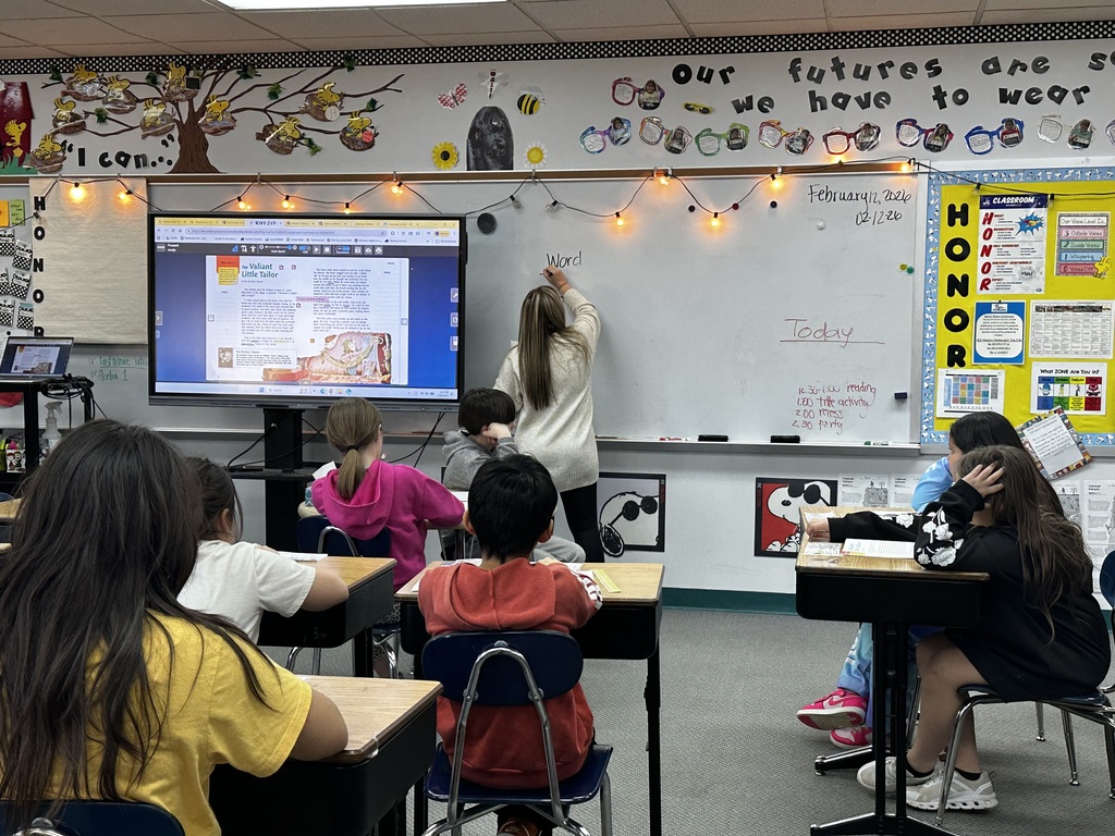 A classroom with children sitting at desks, a teacher writing on a whiteboard, and a monitor on the wall.