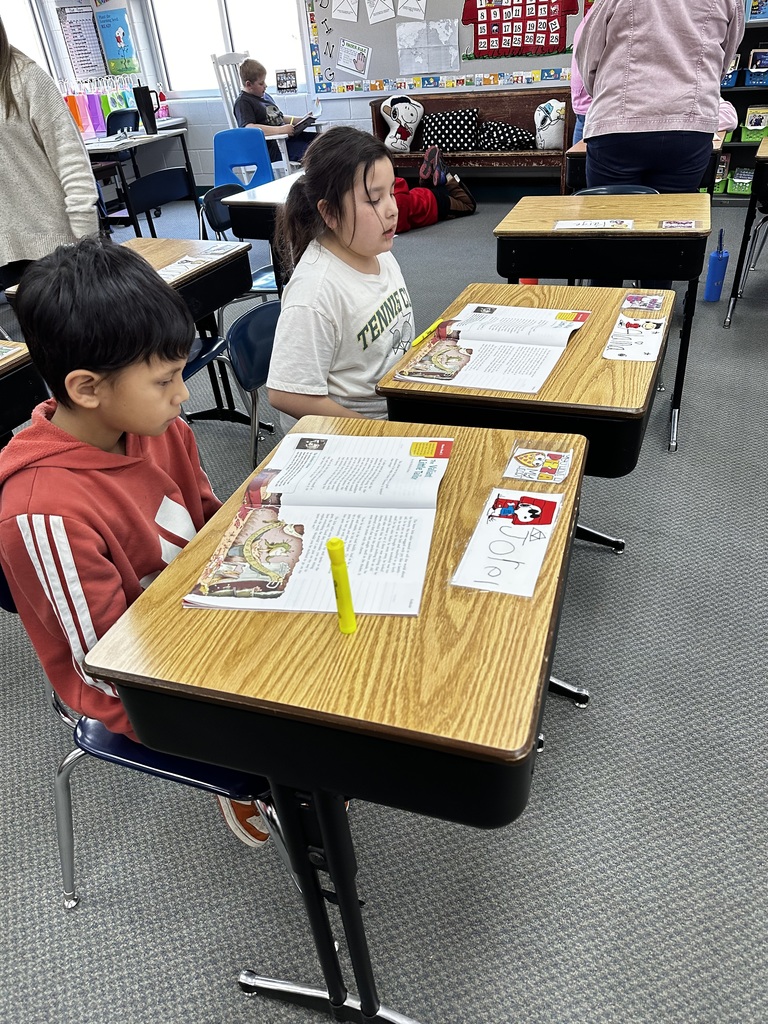 Two children seated at desks, one reading aloud, another listening. Desks are equipped with books and highlighters.