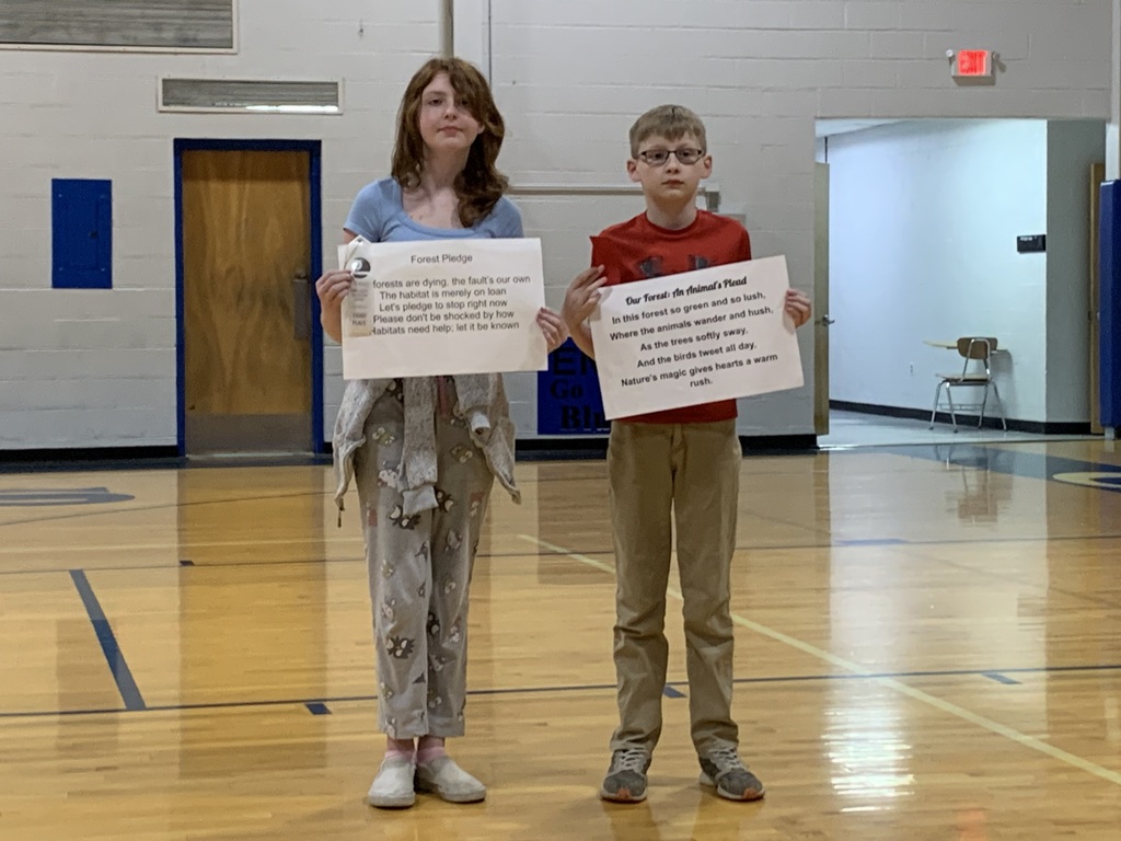 Two students stand in a gym holding signs with writing. One wears a blue shirt, and the other wears a red shirt.