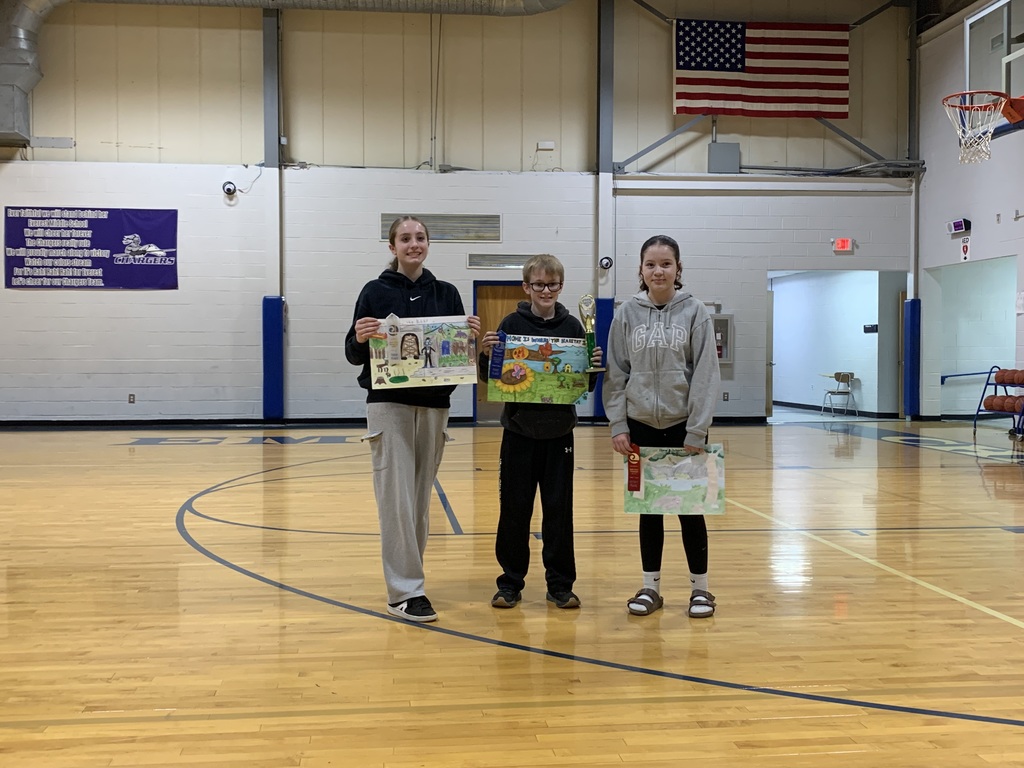 Three students stand in a gym, holding artwork. One displays a colorful poster. Behind them, a basketball hoop and American flag adorn the walls.