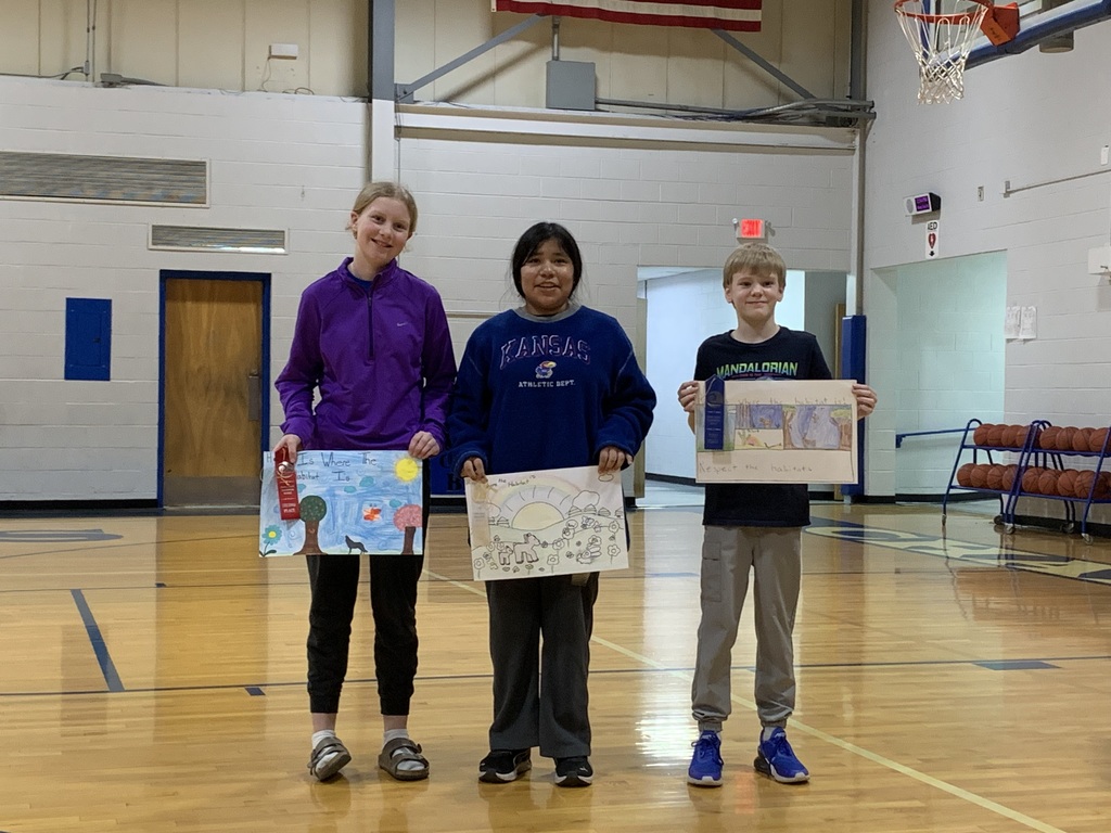 Three children stand in a gym holding artwork. One holds a large drawing. Behind them, a basketball hoop and a flag are visible.