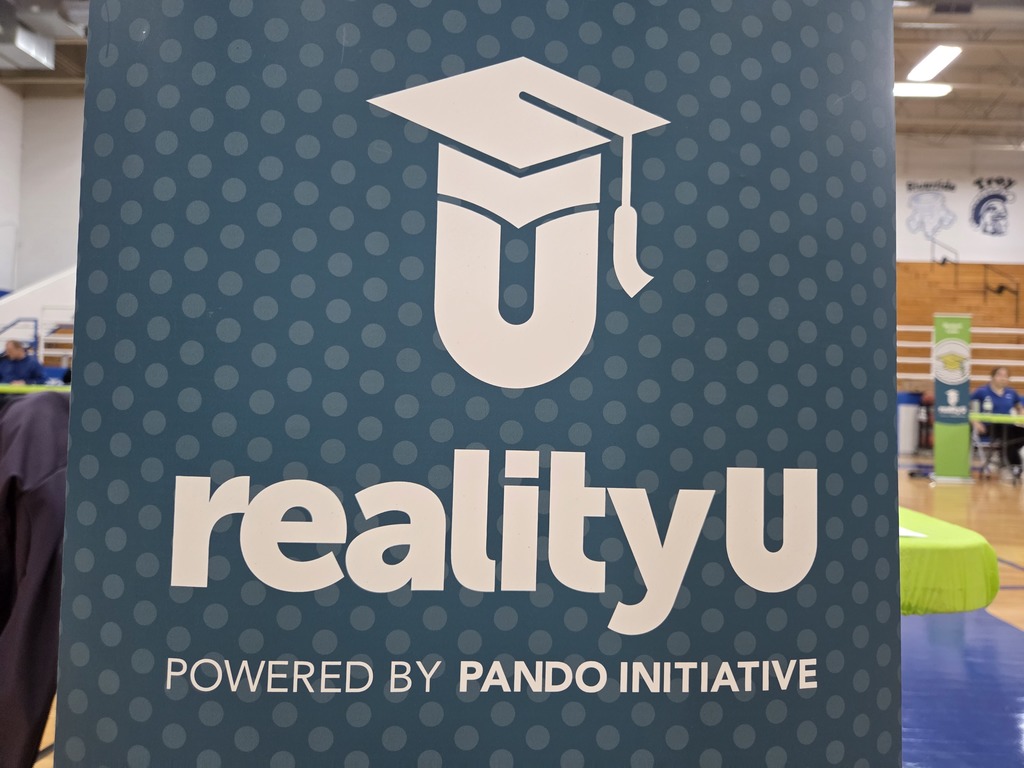 A blue banner with a graduation cap logo, text "realityu" and "POWERED BY PANDO INITIATIVE". Background shows an indoor area with tables and chairs.