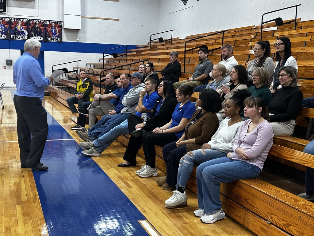 A man speaks to a seated audience in a gymnasium. Audience members wear various colors and styles of clothing.