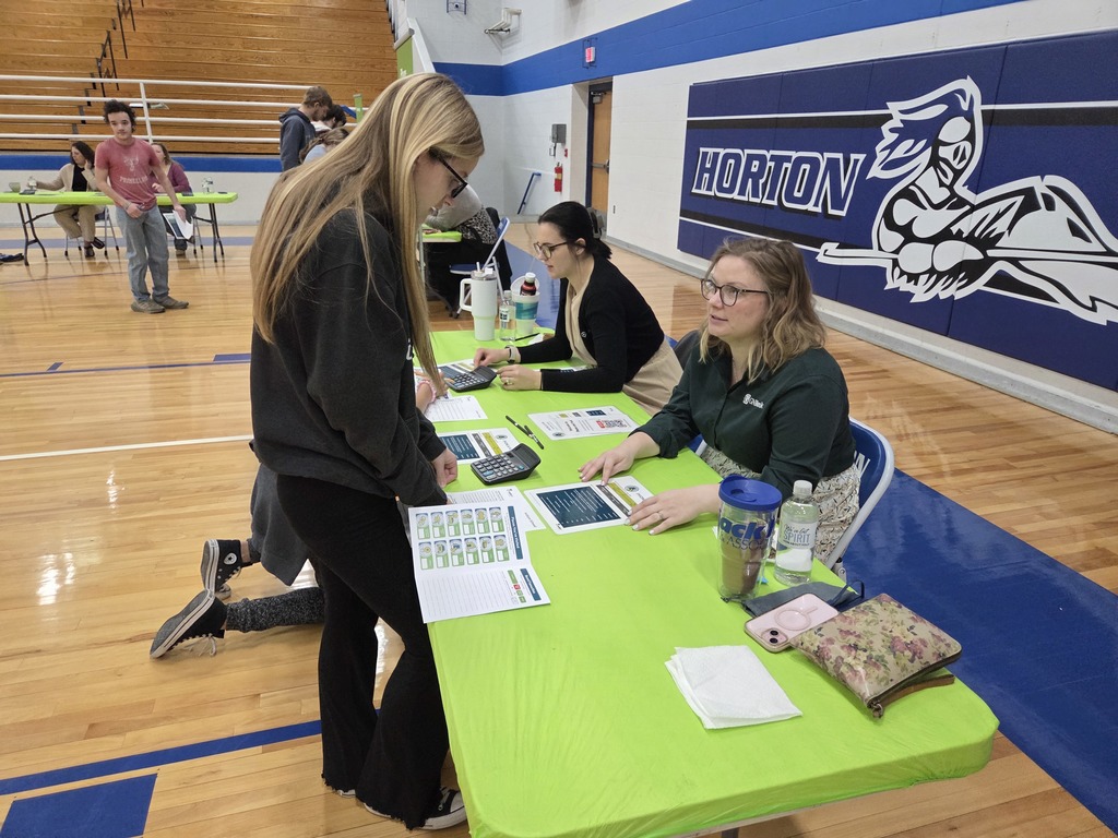 Several people sit at a table in a gymnasium. A woman stands behind the table, looking at papers.