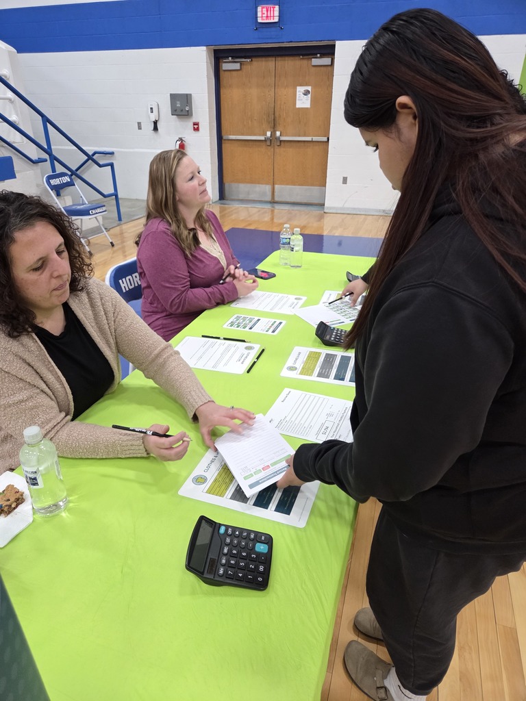 Three people sit at a green table with papers and a calculator. One woman stands and holds papers.
