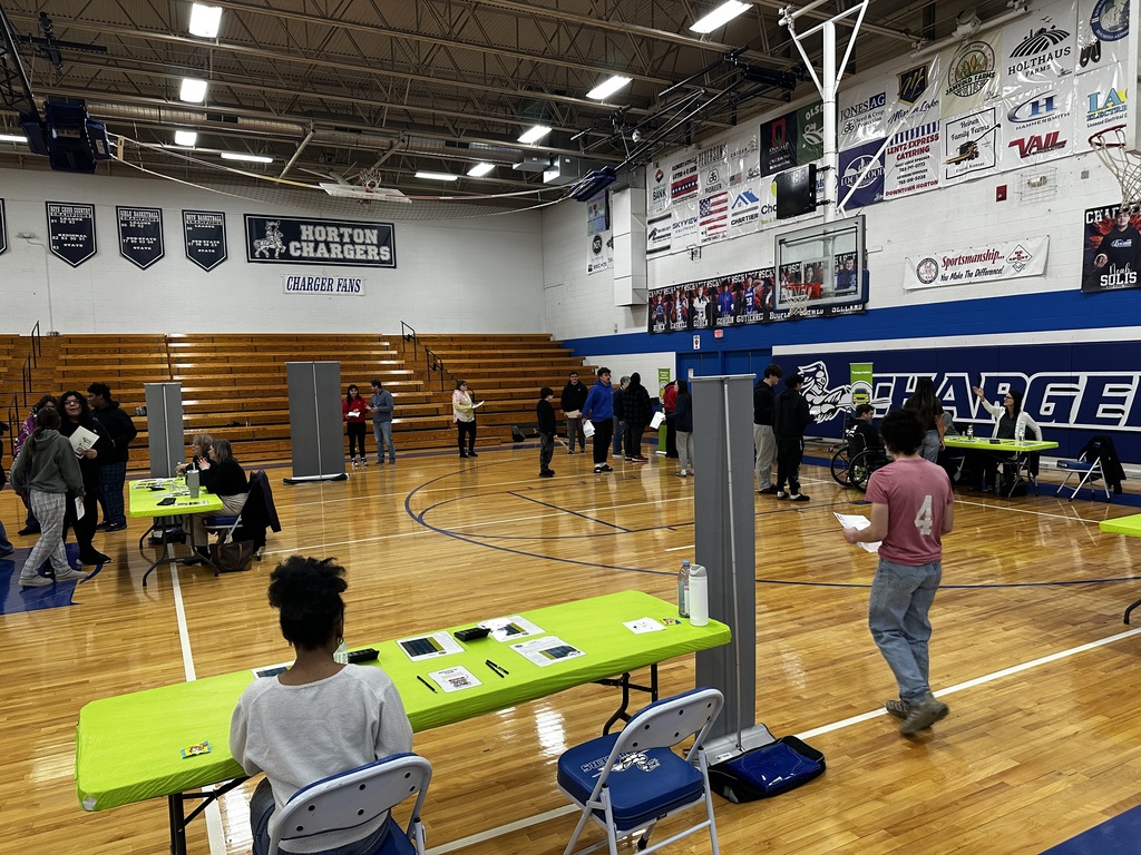 An indoor basketball court with people seated at tables, banners on walls, and a person walking nearby.