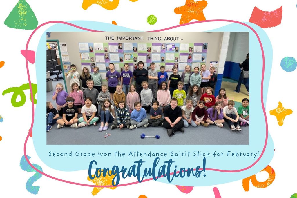 A group of students in a classroom, some sitting on the floor, smiling for a photo. Bright background with decorations. Text reads "Congratulations!" and "Second Grade won the Attendance Spirit Stick for February."