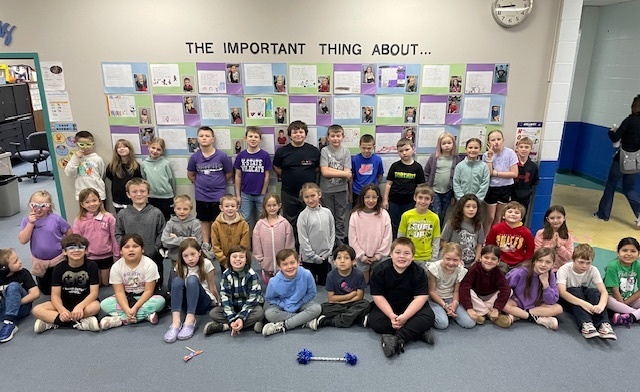 A classroom with students sitting in rows, some standing, with a bulletin board displaying various projects.
