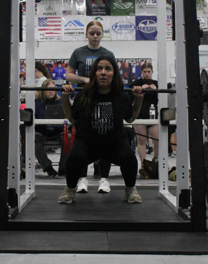 Woman performing a squat with a barbell under a squat rack, with people watching in the background.