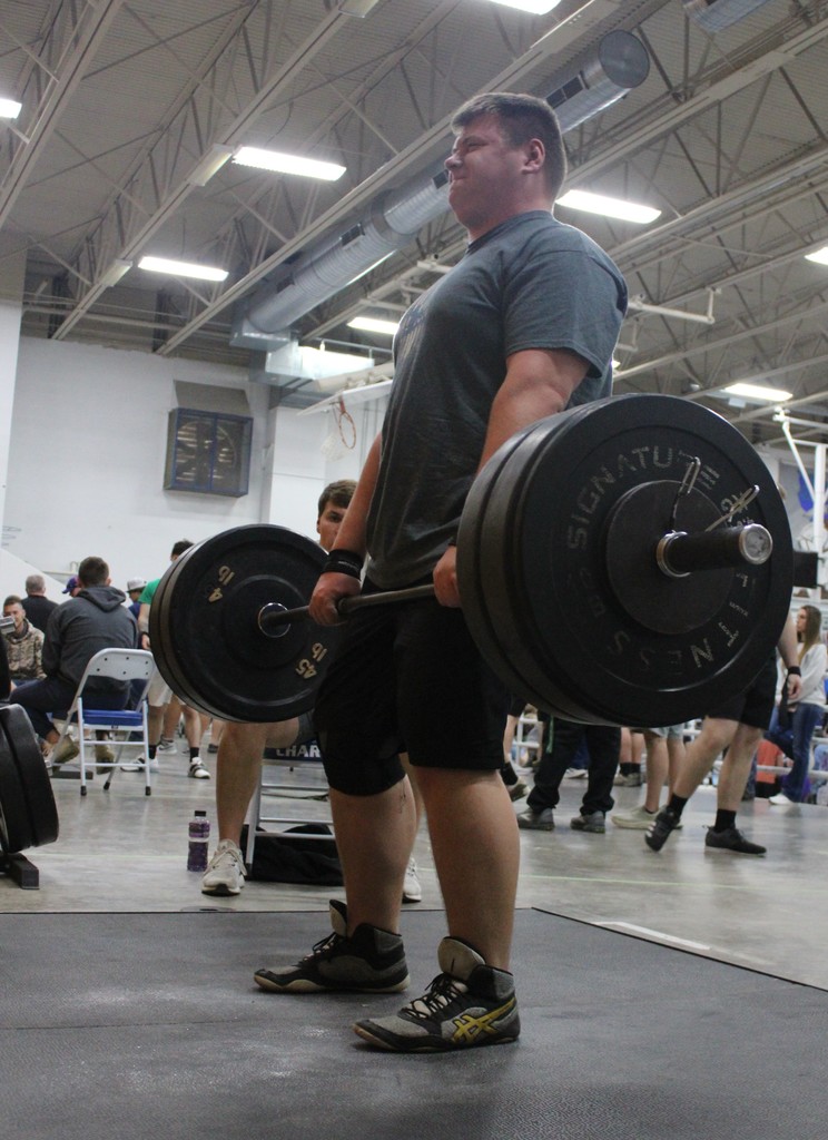 A man lifts a barbell during a workout in a gym with other people in the background.