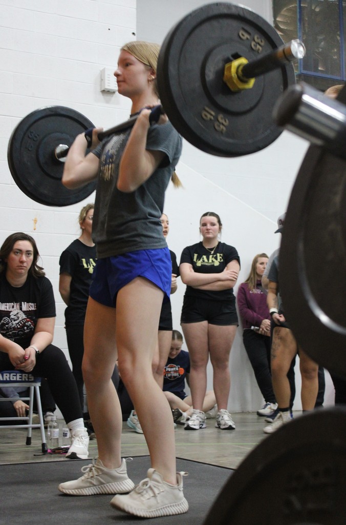 Woman lifting barbell with weights, wearing shorts and sneakers, in gym with several onlookers in background.