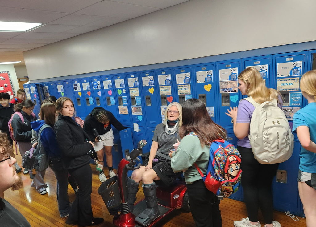 Students at school, some with backpacks, around a person seated on a scooter. Lockers line the wall, adorned with various items.