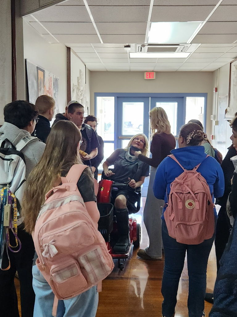 People stand in a hallway, some wearing backpacks. A person in a wheelchair laughs, and a group surrounds them.