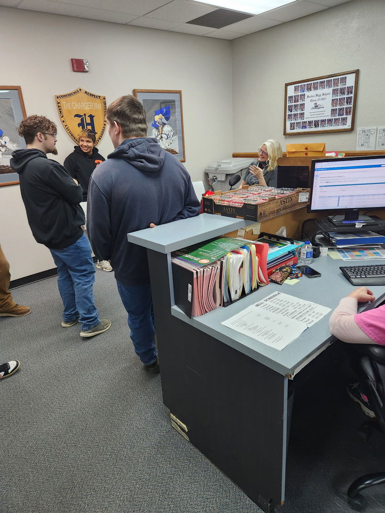 People stand in an office with desks, computers, and frames on walls. A woman speaks on the phone.