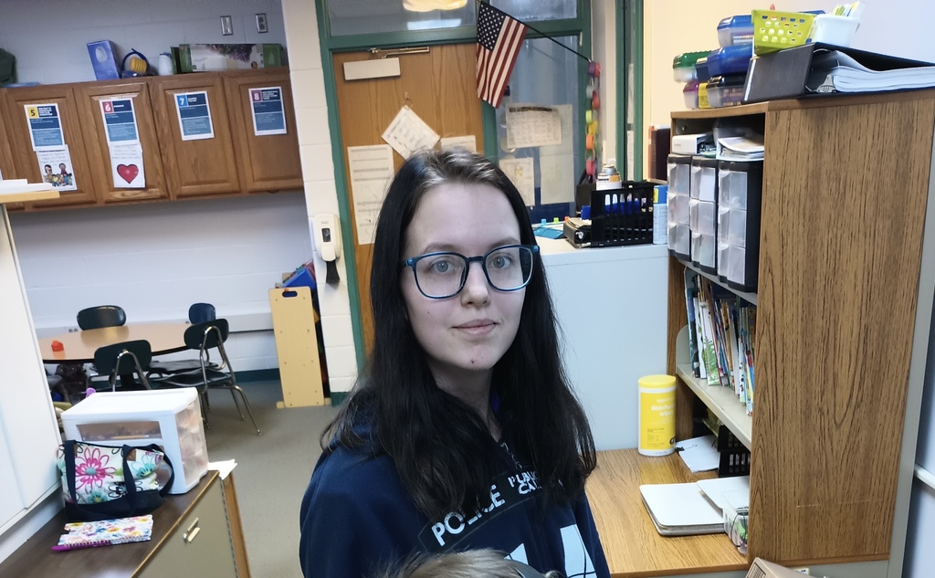A person with glasses and long hair standing in a classroom with a bookshelf and a desk.