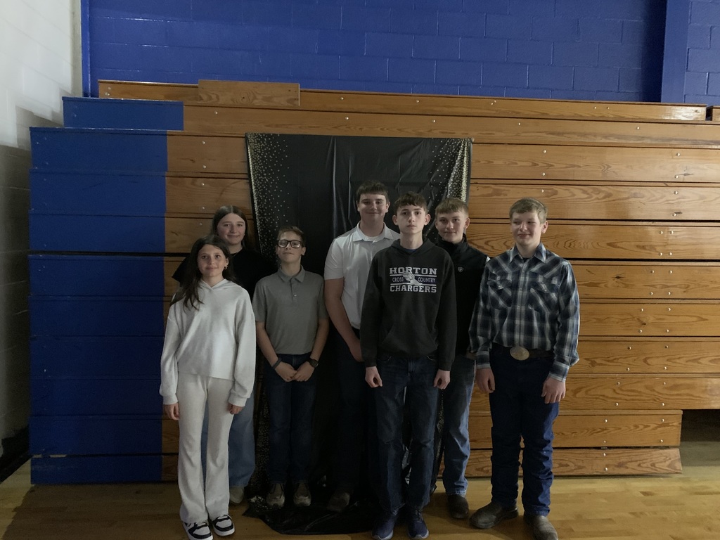 Seven individuals in light clothing stand before a blue wall and wooden bleachers in a sports facility.