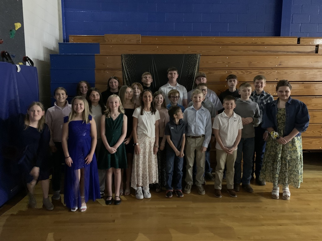 A group of children and adults standing in a room with wooden bleachers in the background.