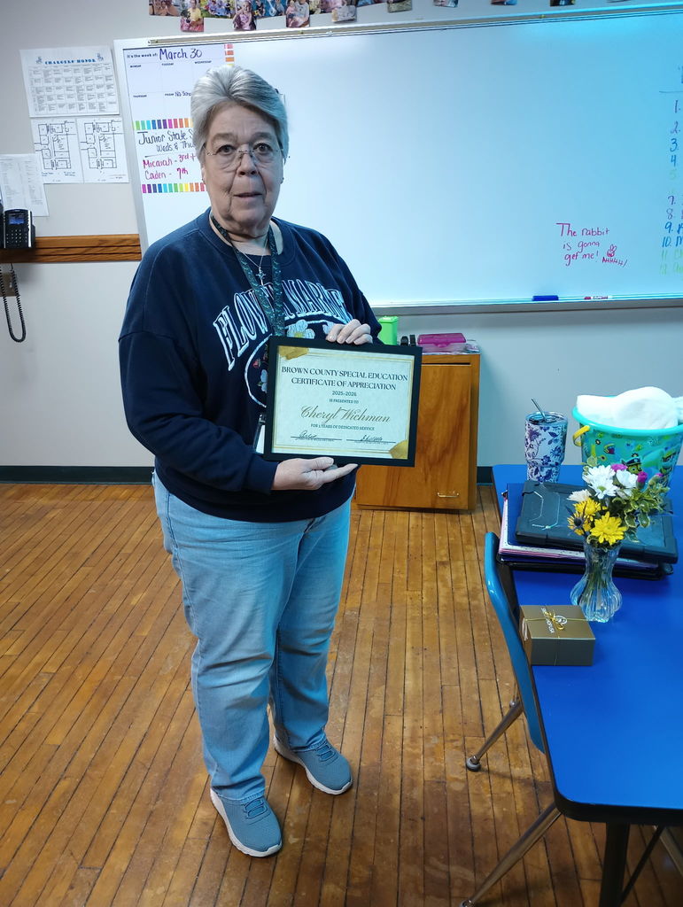 Person with glasses in a classroom holding a framed certificate, standing near a blue table with flowers.