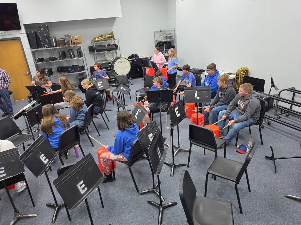 A group of children wearing blue shirts seated in chairs, some holding drumsticks, seated in a room with musical instruments.