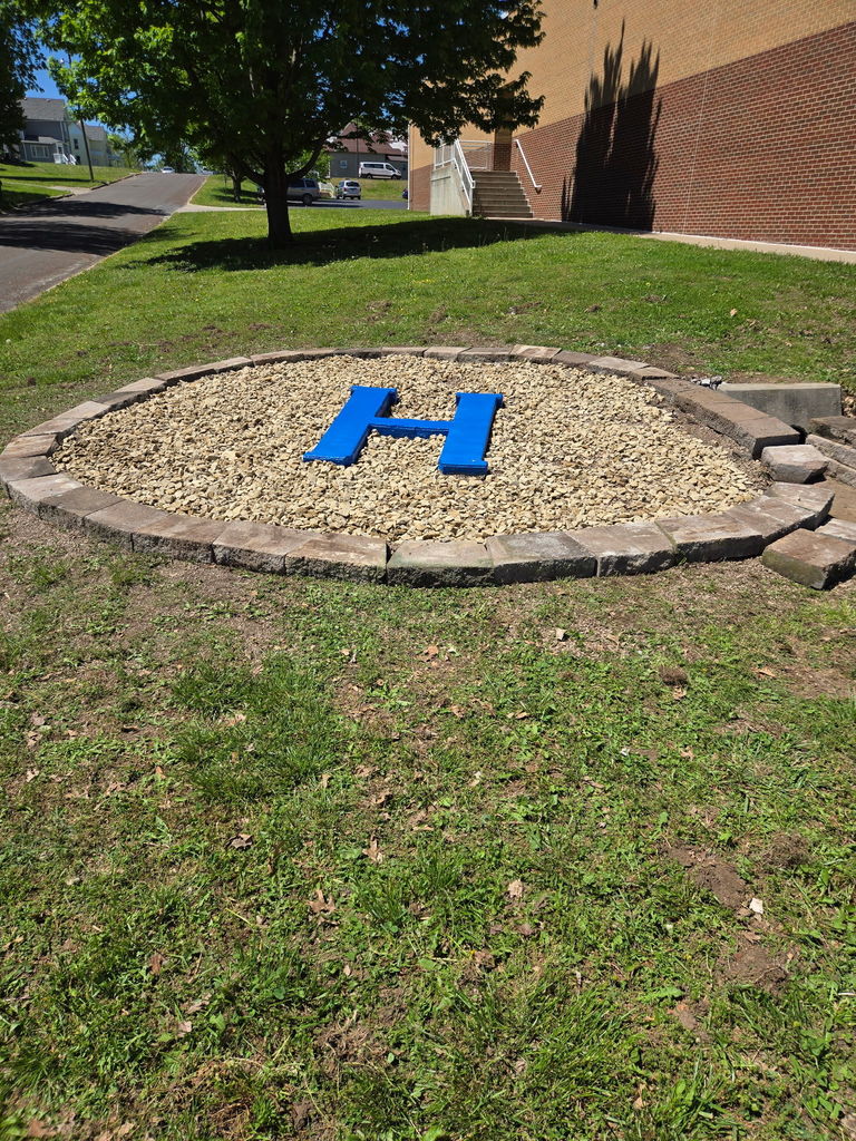 Letter "H" made of blue stones in a grassy area with a tree and building behind.