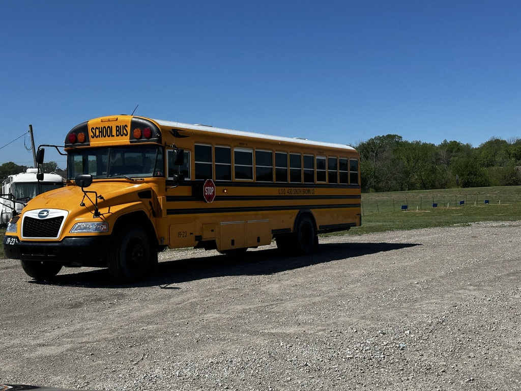 A yellow school bus with 'School Bus' on the side is parked on a gravel road with greenery in the background.
