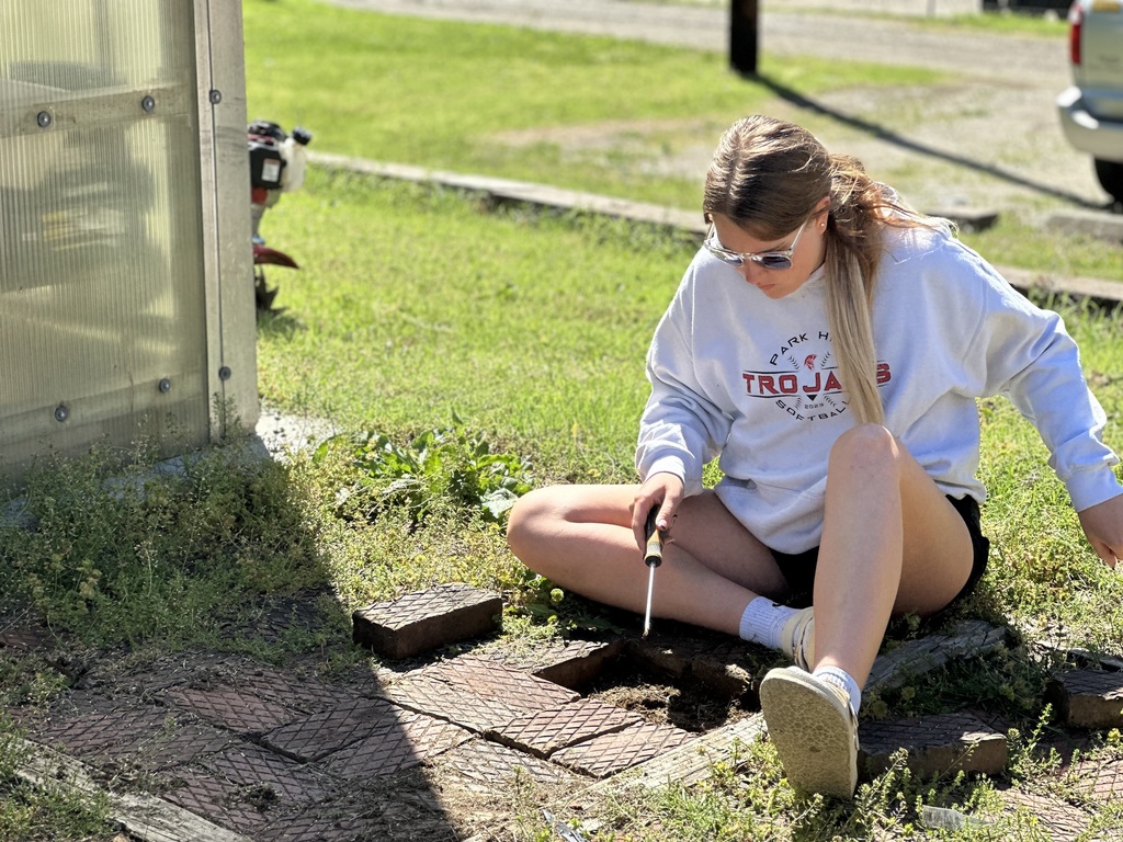 A woman kneels on grass, holding a tool. She wears a white sweatshirt, black shorts, and glasses. Behind her, a structure and vehicle are visible.