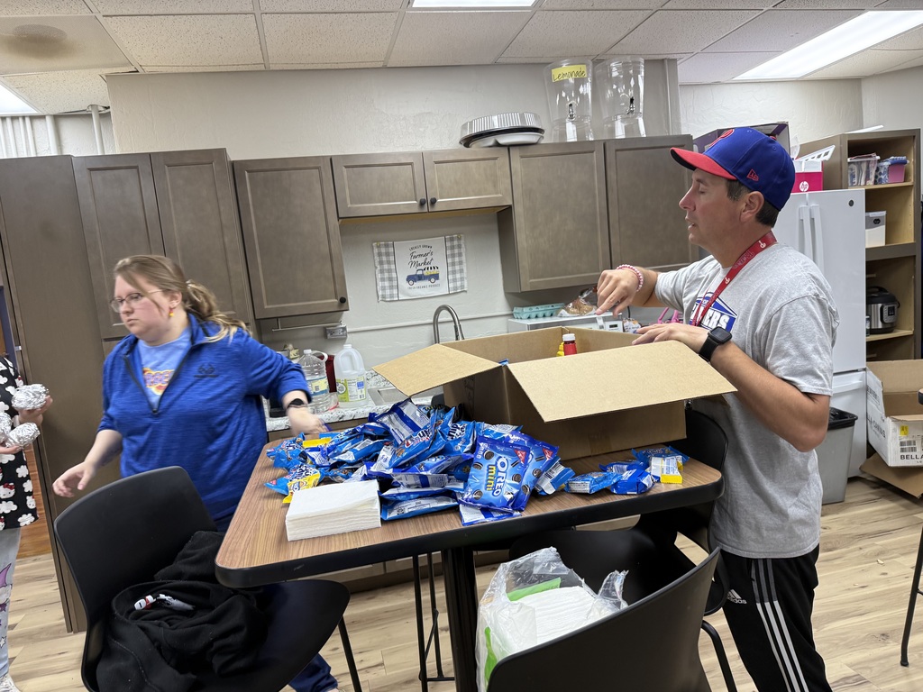 Two people in a kitchen, one with a cap, arranging items on a table. A box is open, revealing multiple items.