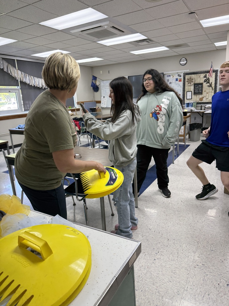 Four individuals in a classroom, two standing at a table, one holding a yellow object, and a fourth running.