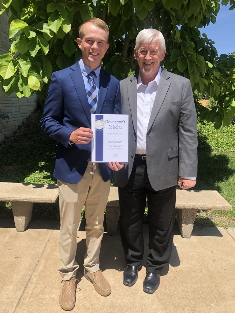 Two men, one holding a certificate, stand in front of a bench and trees. The man holding the certificate wears a suit.