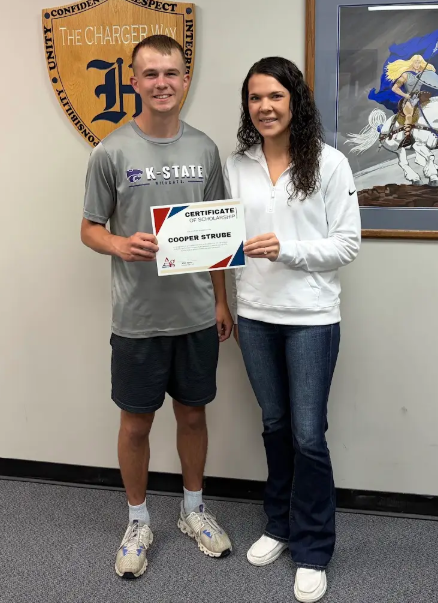 Man and woman holding a certificate standing by a wall with a logo and framed picture.