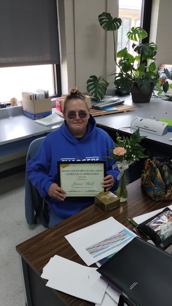 A woman in a blue hoodie holds a framed certificate at a desk with a plant and flowers.