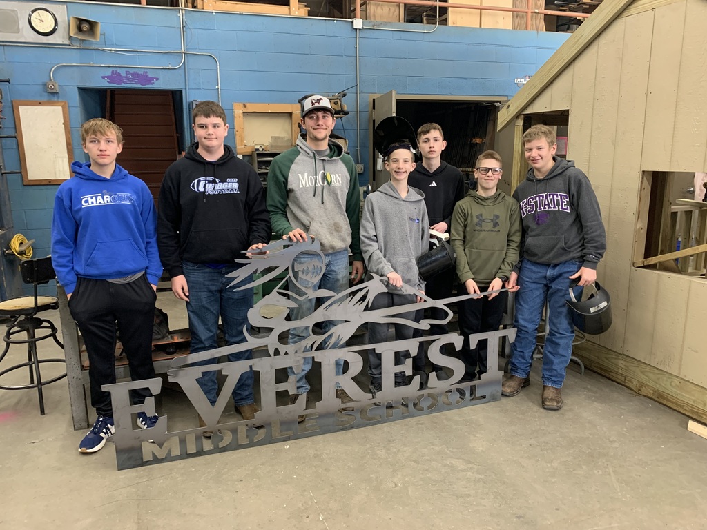 Seven students stand in a room behind a sign that reads "Everest Middle School". They wear hoodies and sneakers.