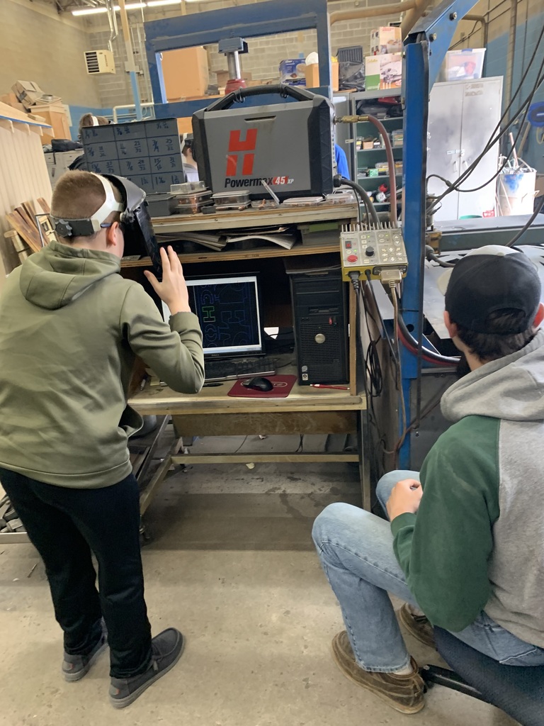 Two people in a workshop. One is using a computer and wearing a virtual reality headset. The other is sitting and watching.