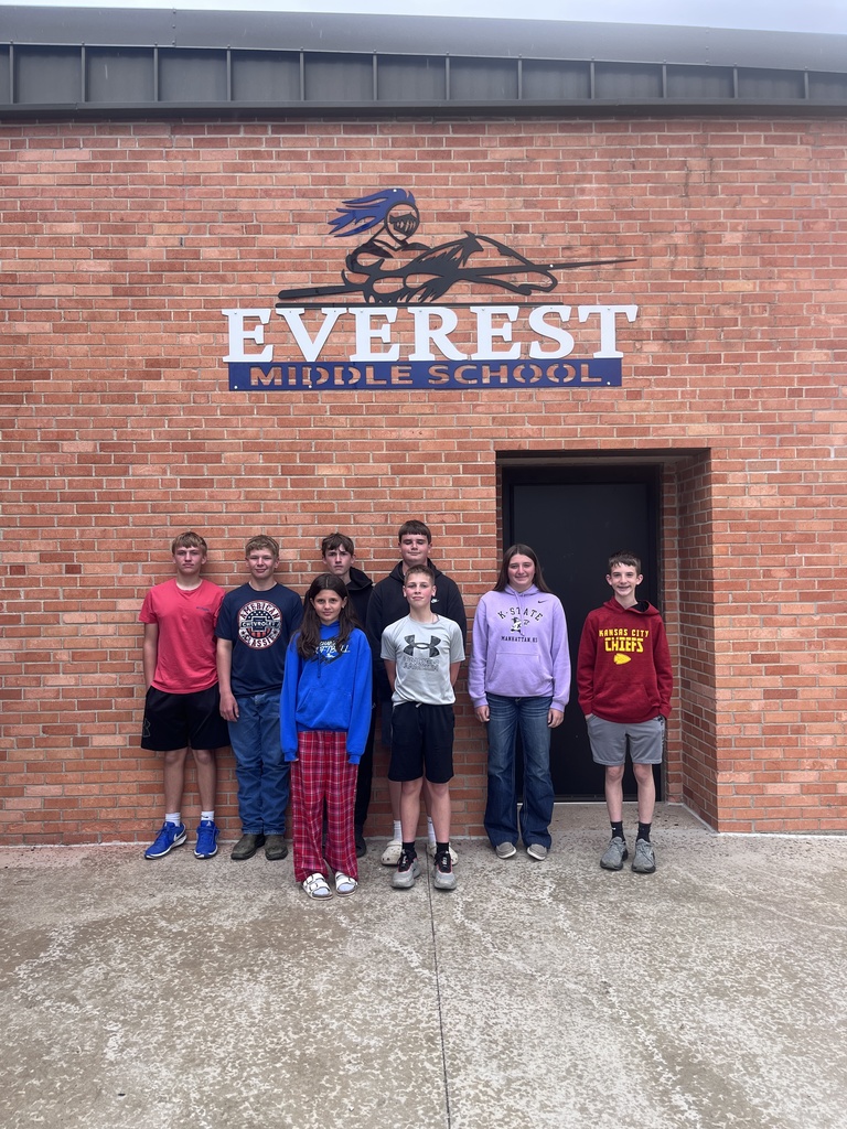 Eight individuals stand before a brick building with a sign reading "Everest Middle School" above the entrance.