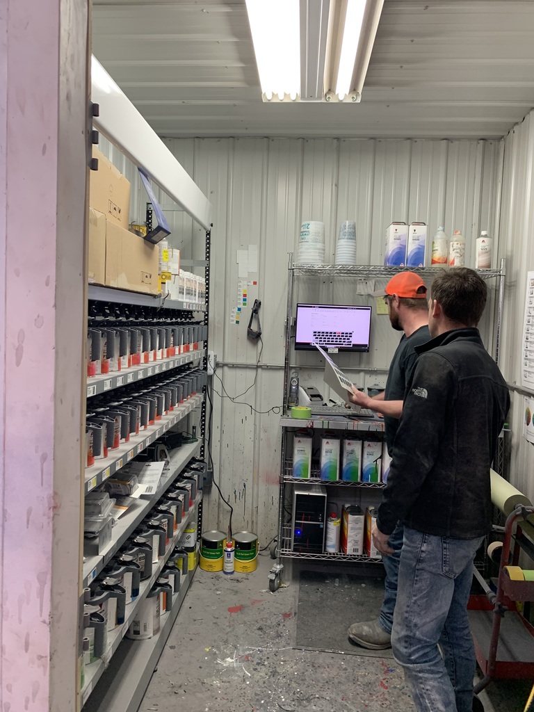Two men in a storage area, one examining a monitor, shelves with boxes and paint cans, fluorescent lighting overhead.