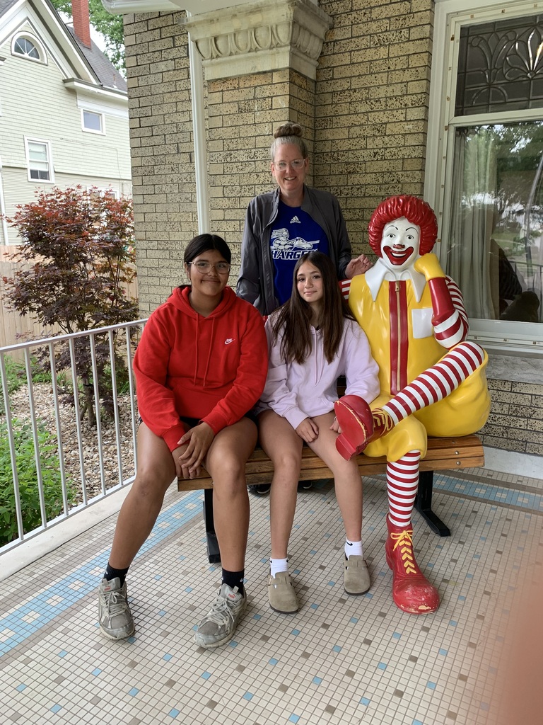 Four people sit on a bench on a porch. Three of them are wearing jackets. A Ronald McDonald statue stands next to them.