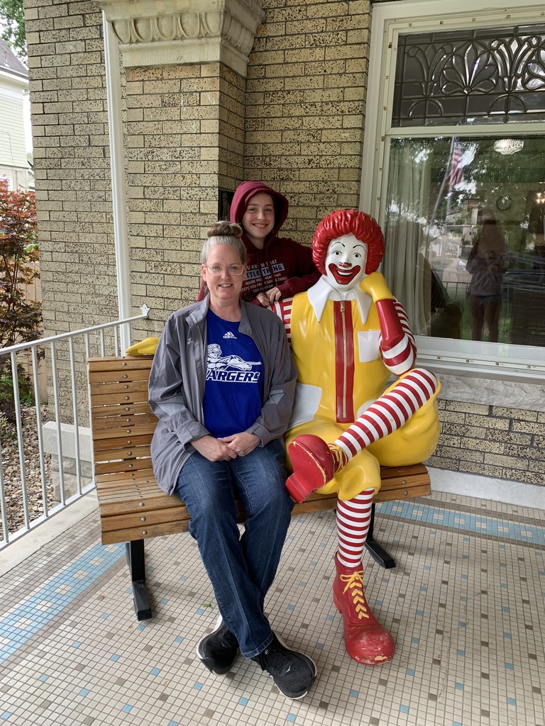 Two people sit on a bench next to a Ronald McDonald statue on a porch. Behind them is a building with a window.