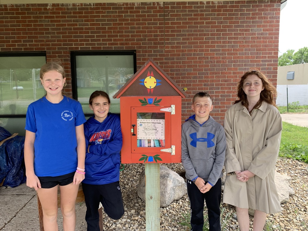 Five children stand by a red birdhouse on a post, in front of a brick building.