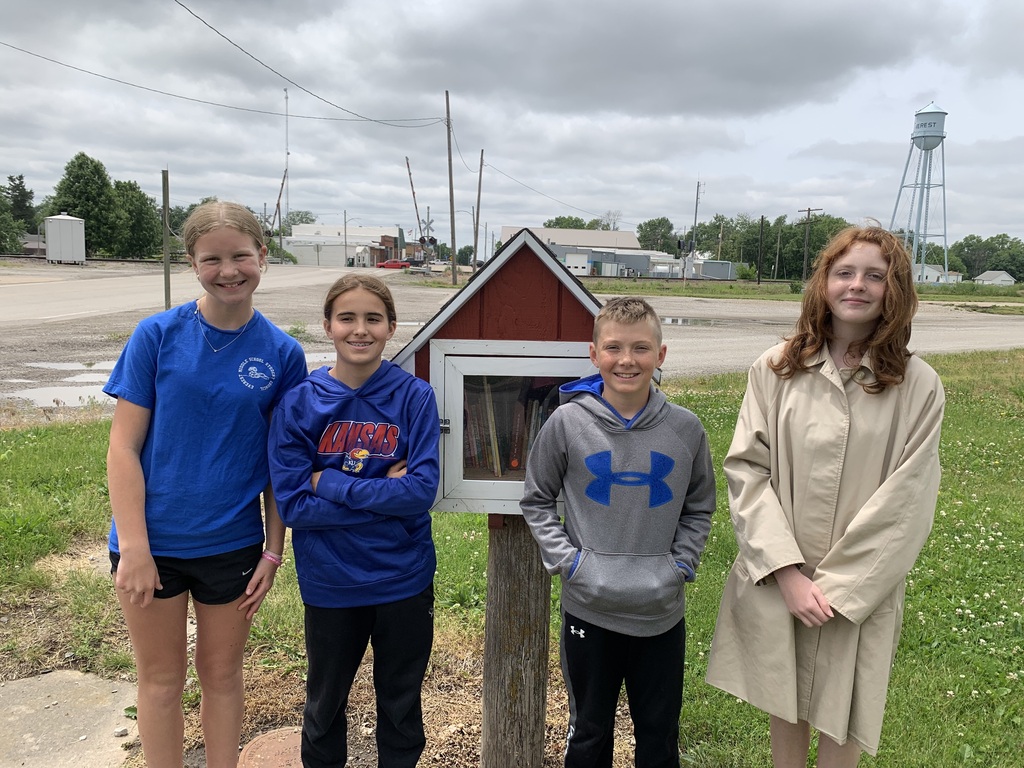 Four kids stand next to a small red box mounted on a wooden post. Behind them is a cloudy sky and some buildings.