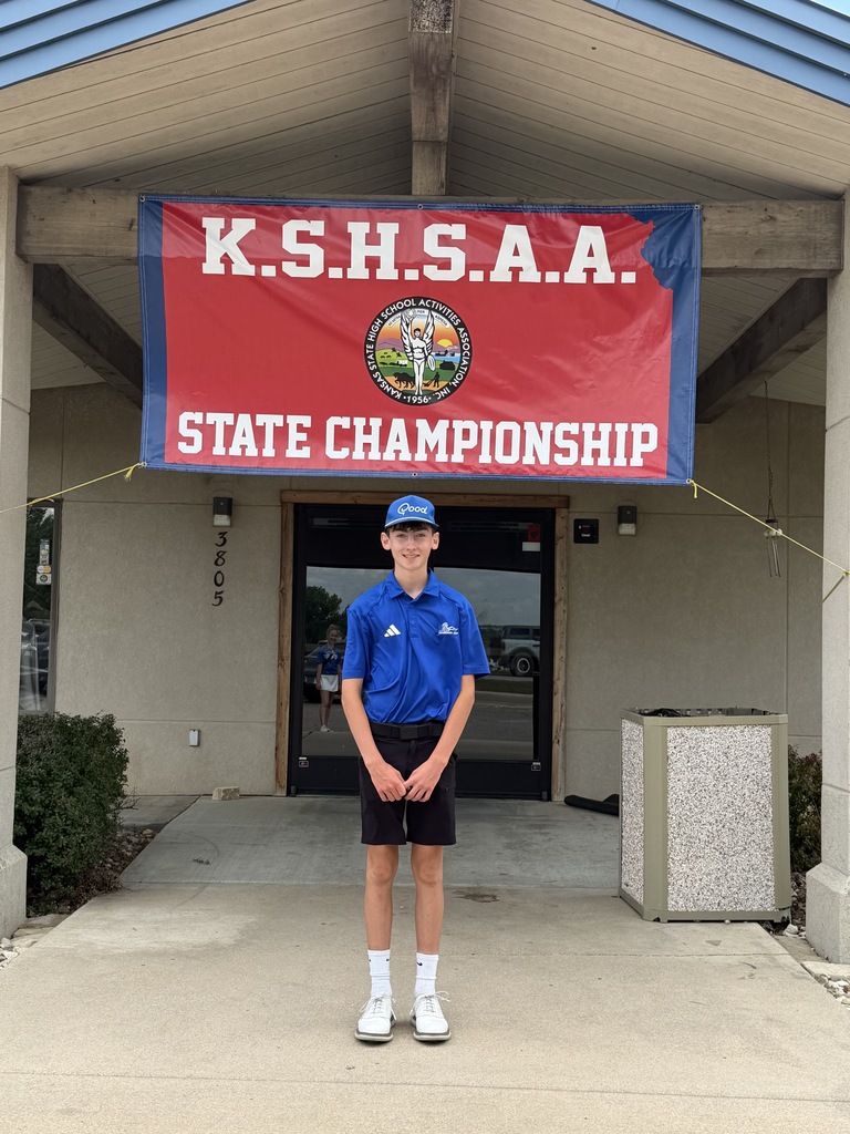 A person in a blue shirt and cap stands in front of a building with a banner reading "K.S.H.S.A.A. STATE CHAMPIONSHIP".