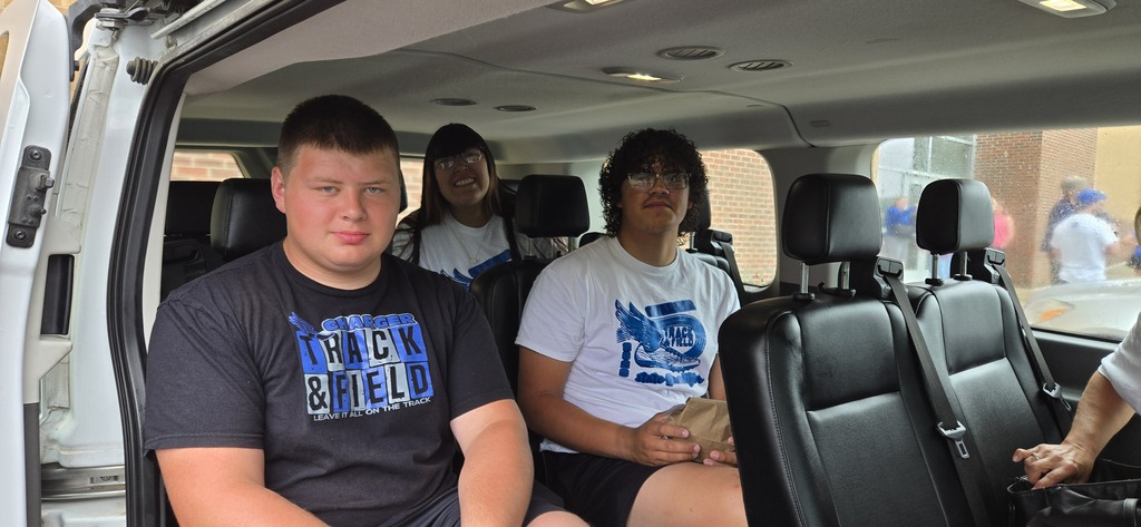 Three individuals sit in a van, two in the front and one in the back. One wears a blue shirt, another a white shirt.
