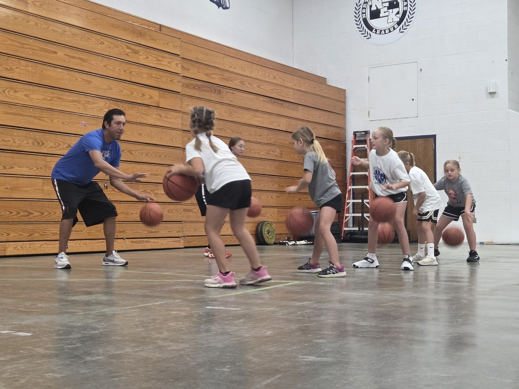 Group of children playing basketball in an indoor gym with wooden wall. A man in blue shirt and black shorts is teaching them.