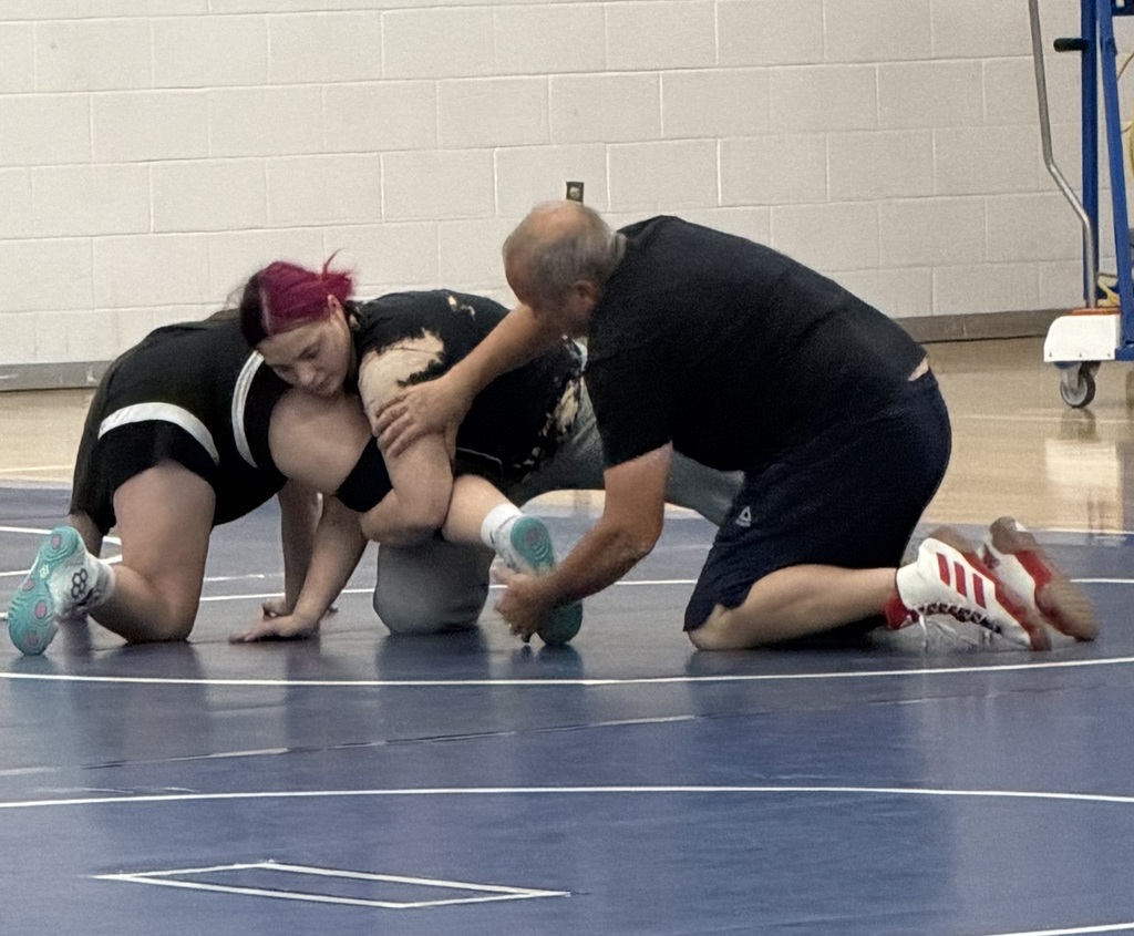 Two people in black uniforms are on a wrestling mat. One is pinned while the other holds them. An adult male referee kneels nearby.
