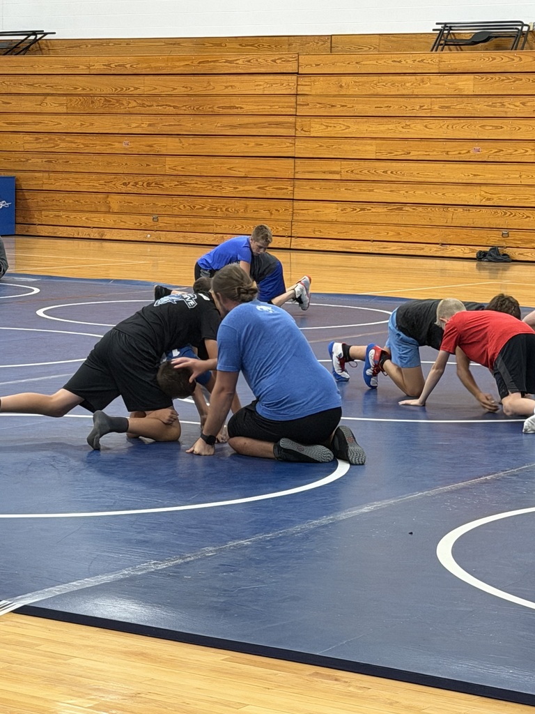 People in various positions on a blue mat inside a gym with a wooden wall.