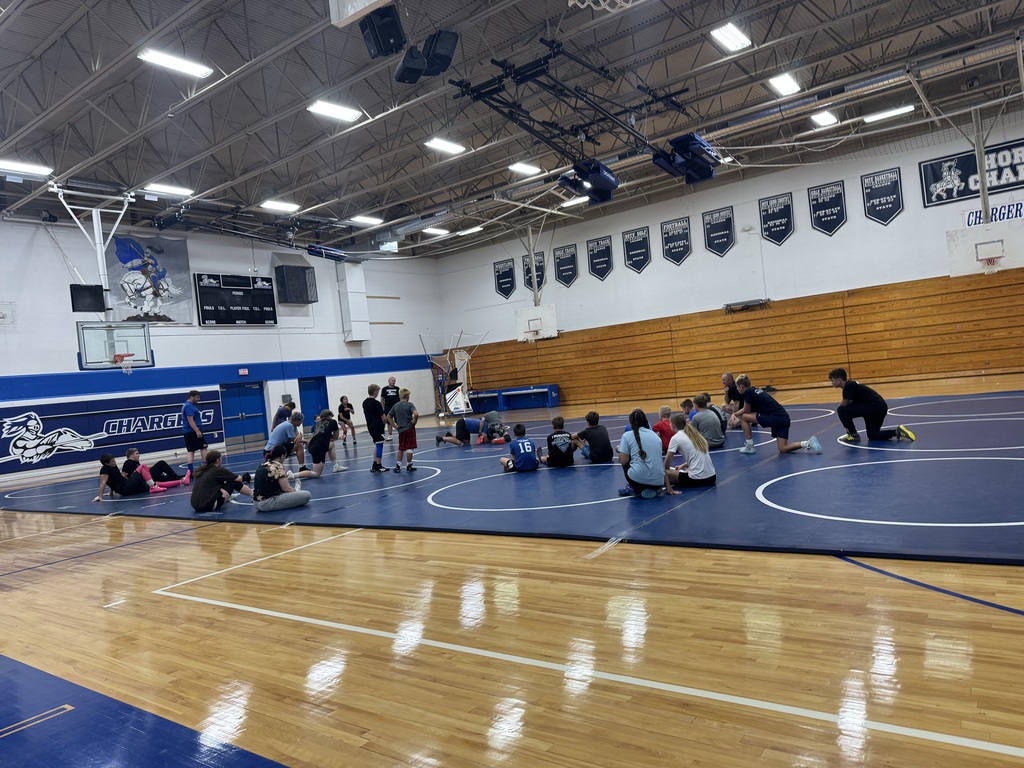 An indoor gym with a blue mat on the floor and people sitting on it, practicing.