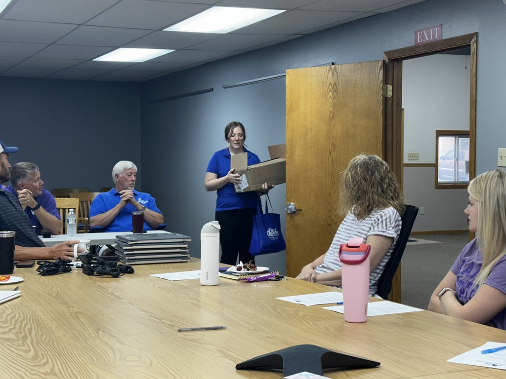 A woman in a blue shirt stands by a door, holding a box, with people seated around a table in a room.